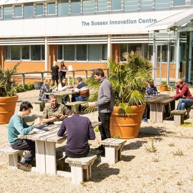 A photograph of the Sussex Innovation Centre in Falmer on a sunny day. Sussex Innovation members are sitting on benches located on the terrace just outside the centre.